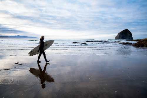 Men moon walks along the smooth shore holding his surfboard, his reflection is seen almost perfectly in the sand below